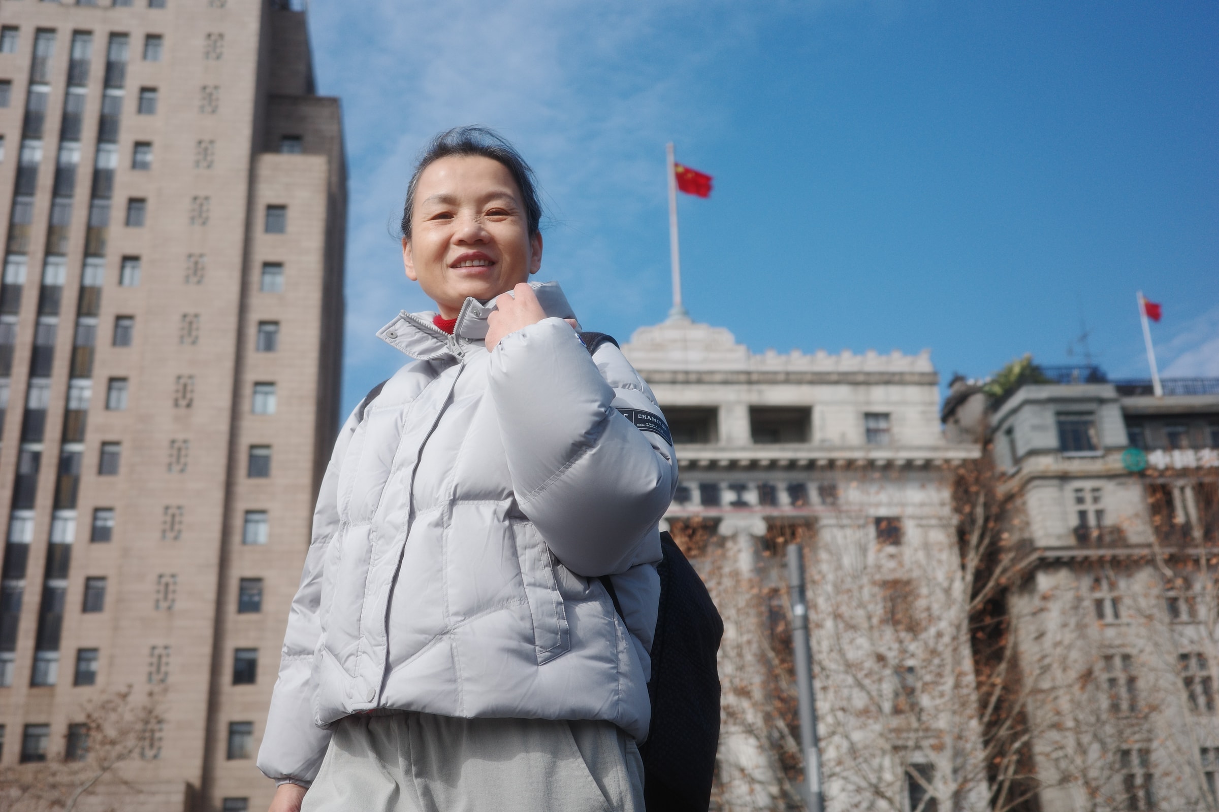 李平身穿浅灰色外套，站在城市建筑前微笑